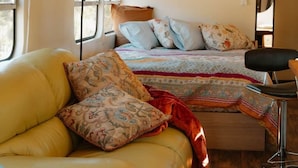 Un intérieur de camping-car compact avec un canapé jaune, un lit avec une literie à motifs et une petite table accompagnée d’une chaise.