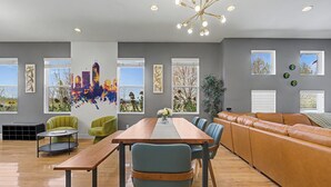 Uma sala de estar moderna com uma mesa de jantar de madeira, cadeiras verdes e azuis, um sofá de couro marrom e grandes janelas com vista para a cidade.