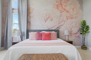 Ein Schlafzimmer mit einer großen floralen Wandmalerei, einem Bett mit rosa und weißer Bettwäsche, zwei Nachttischlampen und einer Topfpflanze.