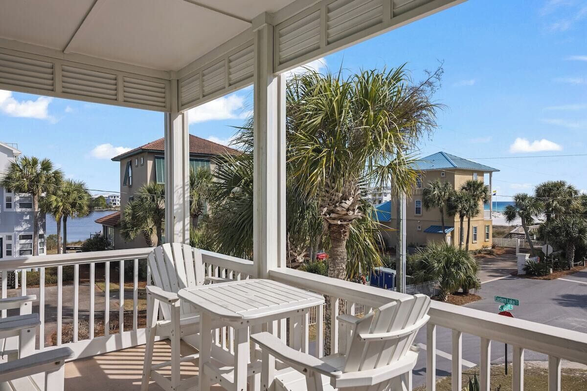 A covered porch with white chairs and a table, overlooking a street with palm trees and houses.