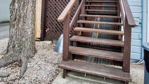 Uma escadaria de madeira com corrimão de metal que leva a um deck, cercada por brita e uma árvore.