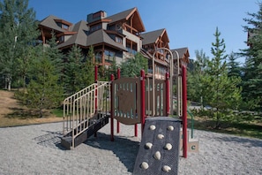 Un parque infantil con tobogán y estructura de escalada, rodeado de árboles y un edificio con techo inclinado.