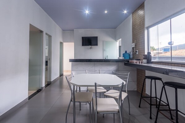 A modern kitchen with a dining area, a TV mounted on the wall, and a large window. - Apartamento Para 6 com Vaga e Churrasqueira (Para de Minas)