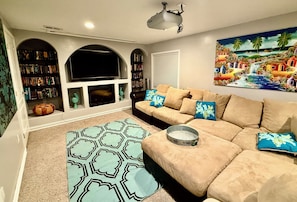 A living room with a beige sofa, a flat-screen TV, a bookshelf, and a painting on the wall. - Spacious 4-bedroom house in beautiful West Jordan with AC and gym (West Jordan)