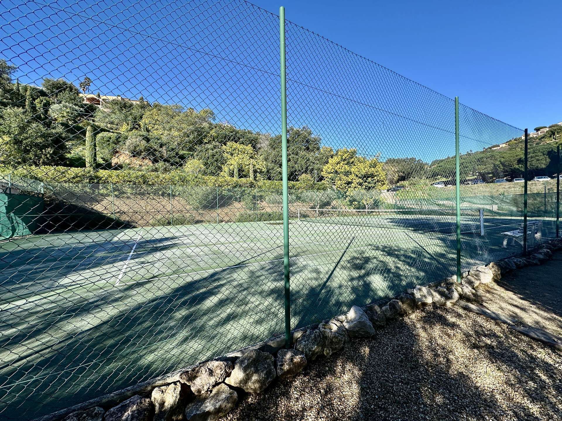 Cancha de tenis rodeada por una cerca de alambre con un borde rocoso y árboles al fondo.