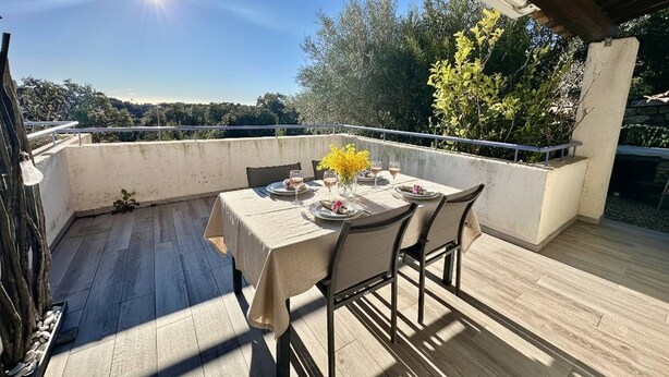 Un área de comedor al aire libre con una mesa dispuesta para cuatro personas, rodeada de sillas, sobre una terraza de madera con vistas a árboles y colinas.