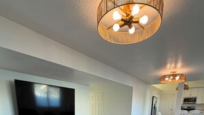 Un salón moderno con televisor de pantalla plana, sofás grises, suelo de madera y una zona de cocina al fondo.