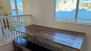 Una habitación con una amplia mesa de madera, un banco, una barandilla blanca y un ventanal con vistas a edificios y montañas.