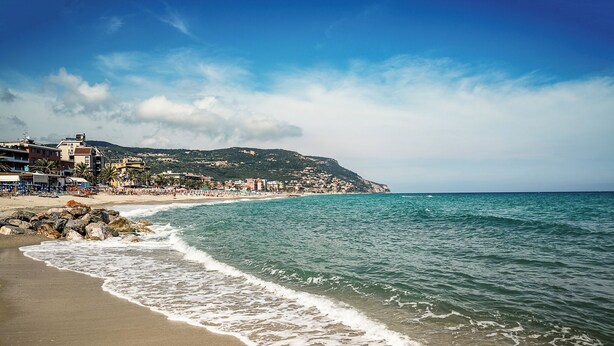 Una spiaggia con un litorale roccioso, cielo sereno e edifici sullo sfondo.