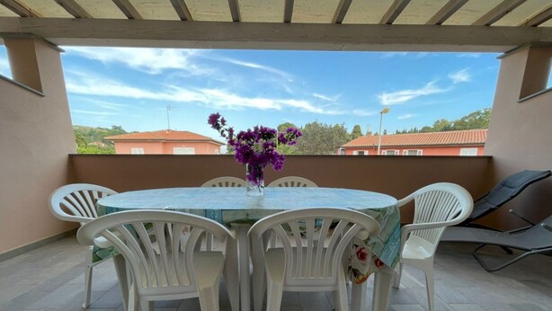 Une terrasse couverte avec une table ronde, des chaises blanches, un vase rempli de fleurs violettes et, en arrière-plan, une vue sur des bâtiments.