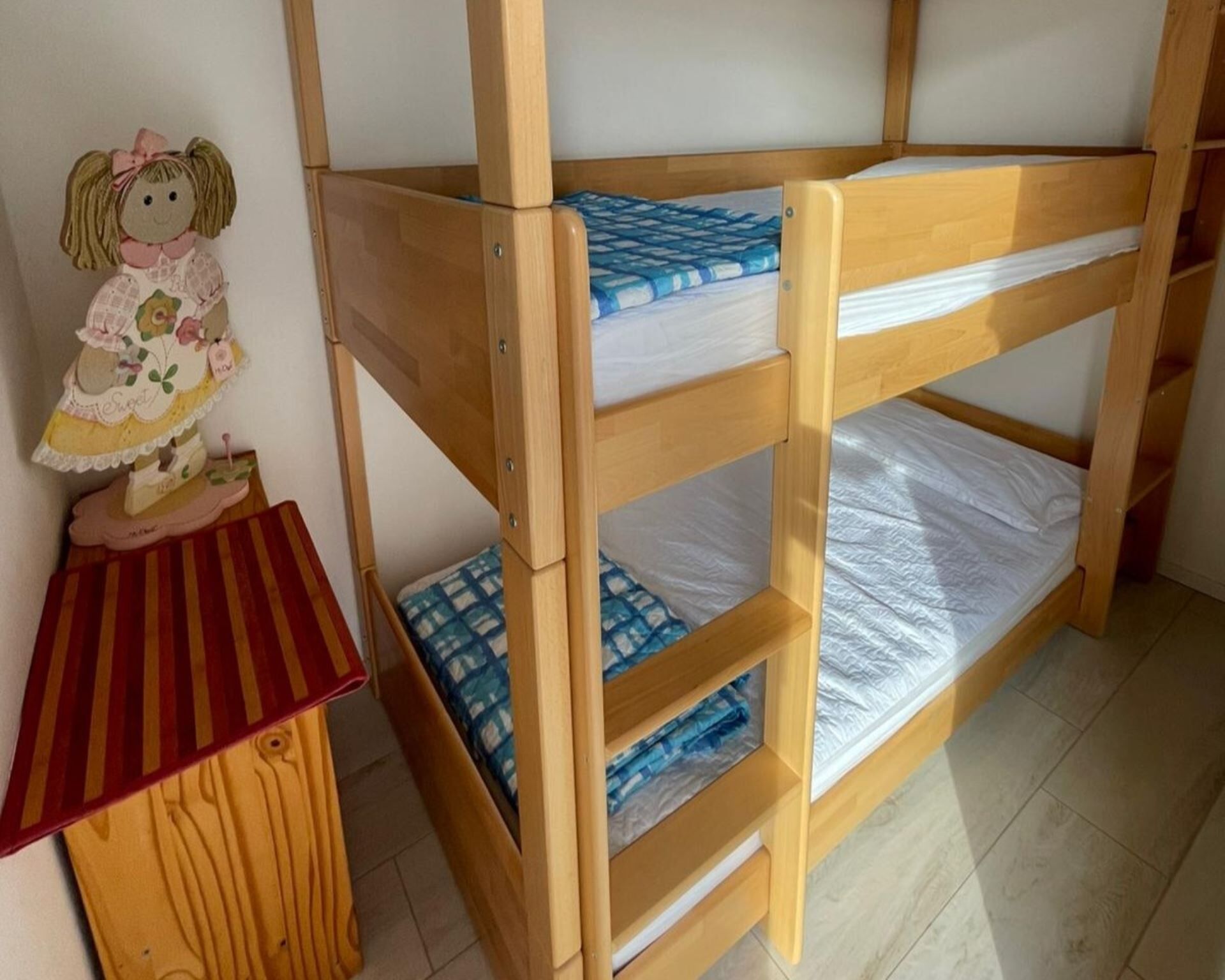 A bunk bed with a wooden frame, a small table with a striped cloth, and a doll on the table.