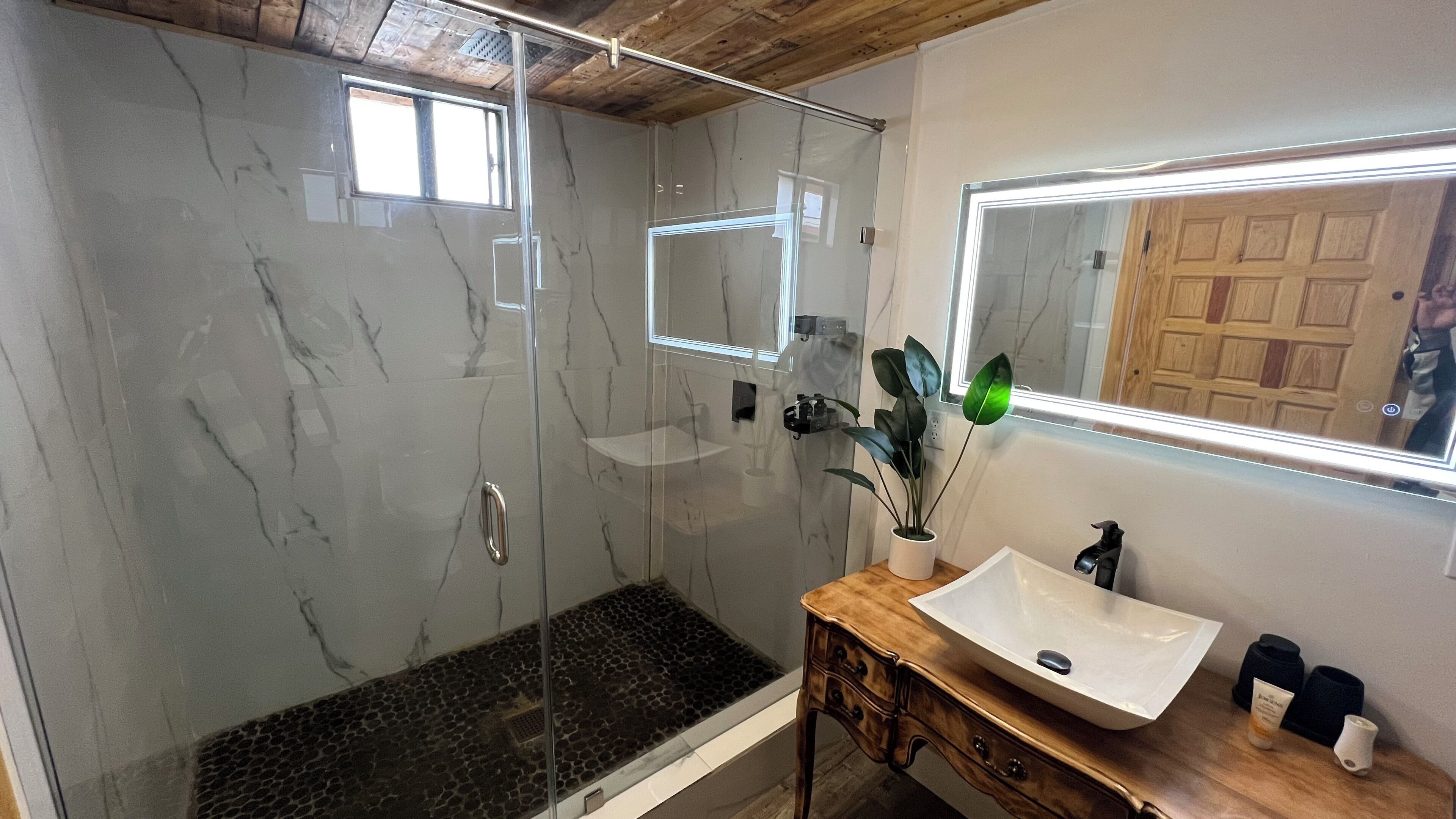 Un baño con ducha acristalada, tocador de madera con lavabo blanco y espejo sobre el tocador.