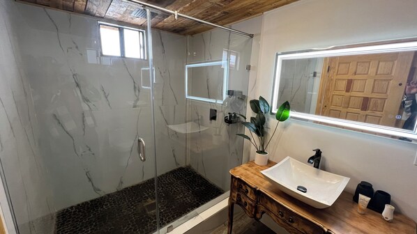 A bathroom with a glass-enclosed shower, a wooden vanity with a white sink, and a mirror above the vanity. - Hacienda 7 Amores (Valle de Guadalupe)