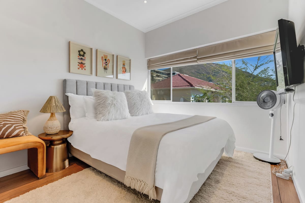 A bedroom with a bed, bedside table, lamp, artwork, window, and fan.