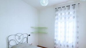Een kleine, nette kamer met een eenpersoonsbed, een groen plankje en een raam met een dunne gordijn.
