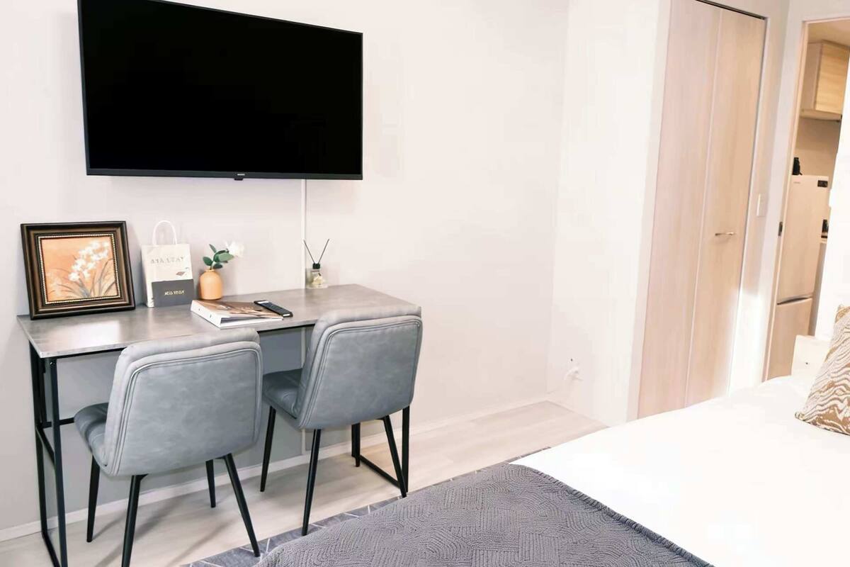 Une chambre d’hôtel moderne dotée d’un téléviseur à écran plat fixé au mur, d’un bureau avec deux chaises, d’un lit agrémenté d’un coussin à motifs et d’une porte donnant sur une autre pièce.
