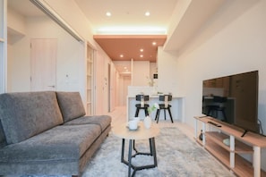 Un salon moderne avec un canapé, une table basse ronde, un téléviseur posé sur un meuble en bois, et un coin cuisine à l’arrière-plan.