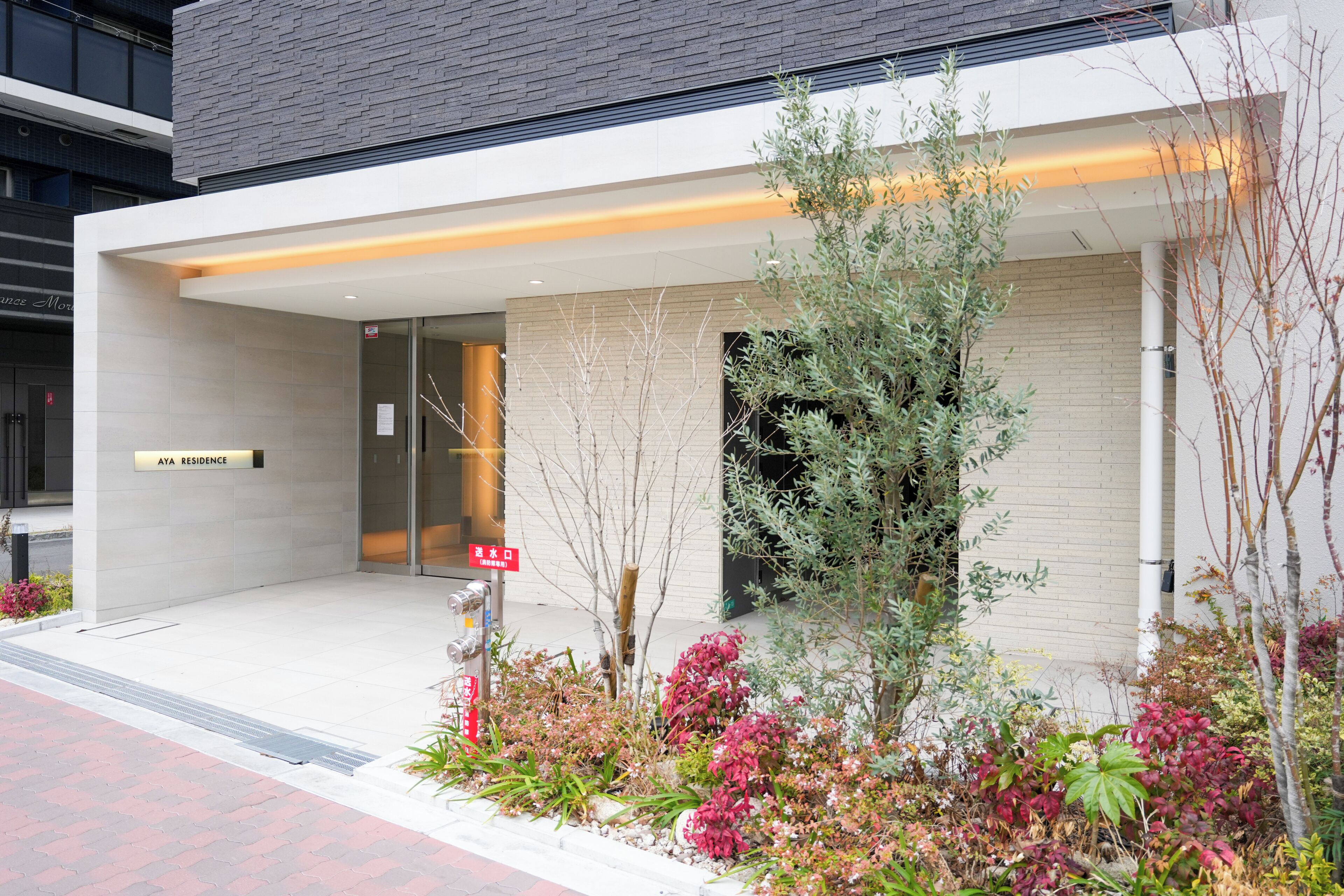 A modern building with a sign reading 'AYA RESIDENCE,' a glass entrance, and a landscaped garden with various plants.