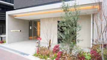 A modern building with a sign reading 'AYA RESIDENCE,' a glass entrance, and a landscaped garden with various plants.