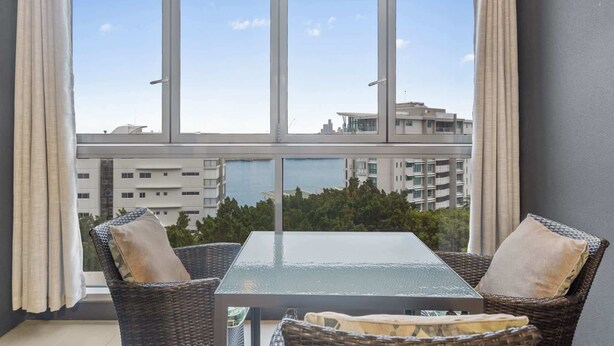 A balcony with wicker chairs and a glass table, overlooking a cityscape and water. - Hamilton Riverview Apt | Balcony & Pool (Hamilton)