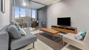 A modern living room with a sofa, a smaller couch, a TV, and a sliding glass door leading to an outdoor balcony. - Hamilton Riverview Apt | Balcony & Pool (Hamilton)