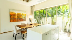 Una cocina moderna con encimera blanca, una mesa de comedor de madera y sillas. Hay un ventanal con vistas a zonas verdes.