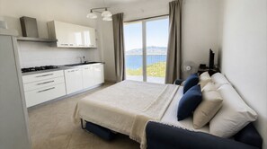A bedroom with a bed, pillows, a view of the outdoors, a kitchen with cabinets, and a fan. - Residenza Bellavista - Bianco (Capoliveri)