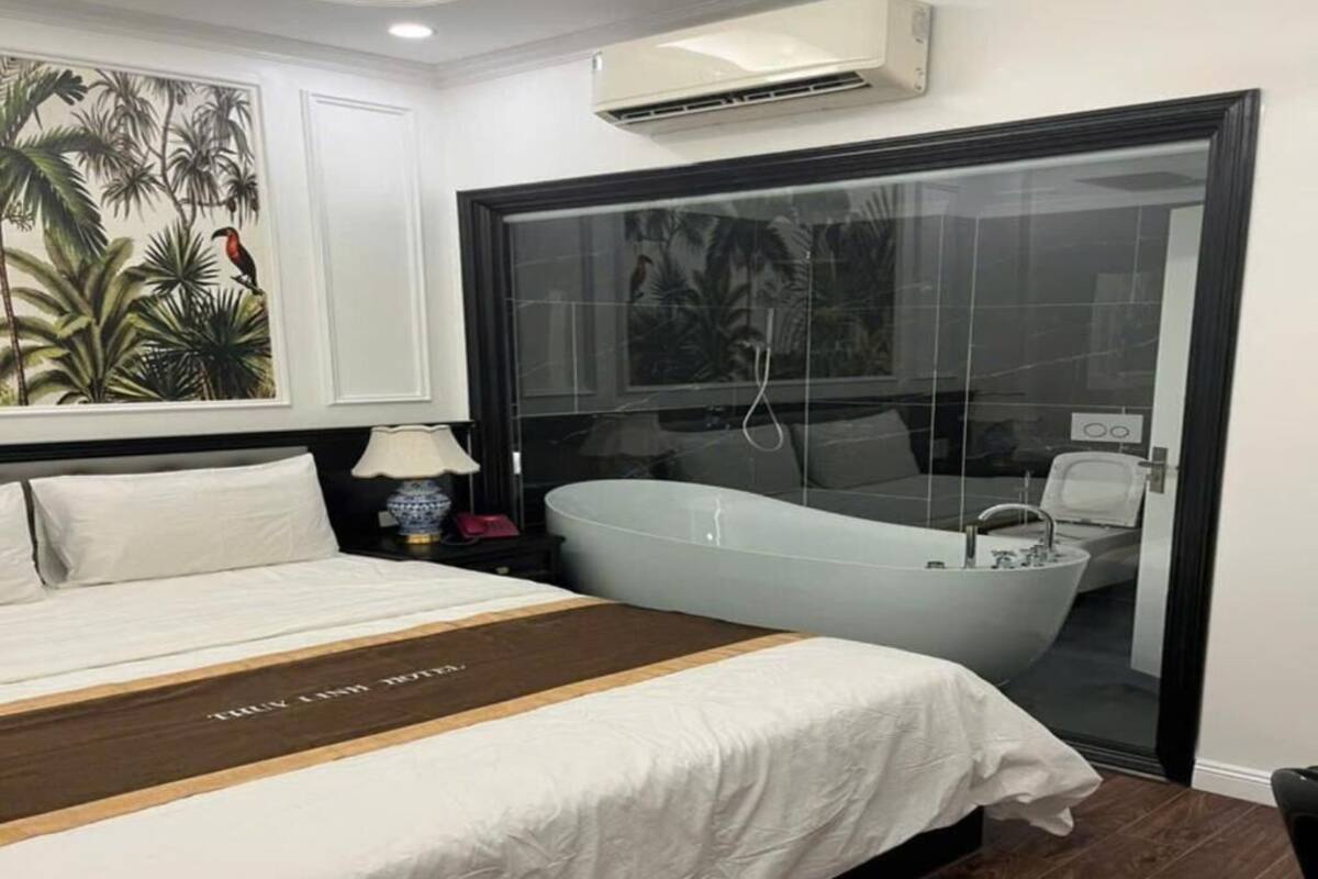 A bedroom with a large bed, a nightstand, a lamp, and a bathtub visible through a glass door.