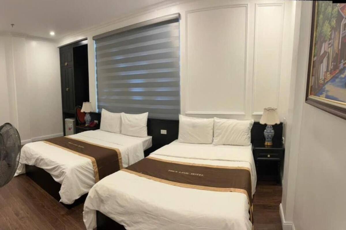 A hotel room with two beds, a fan, a nightstand, and a painting on the wall.