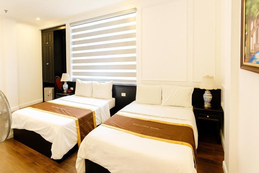 A hotel room with two beds, a large window with blinds, a bedside table with a lamp, and a fan.