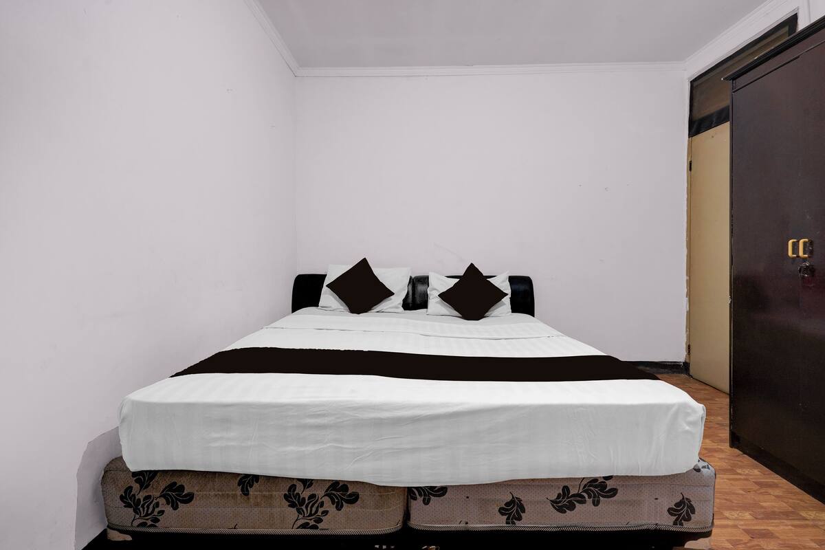 Una cama individual con una colcha blanca y marrón, dos almohadas negras y un cabecero oscuro en una habitación con paredes blancas y suelo de madera.