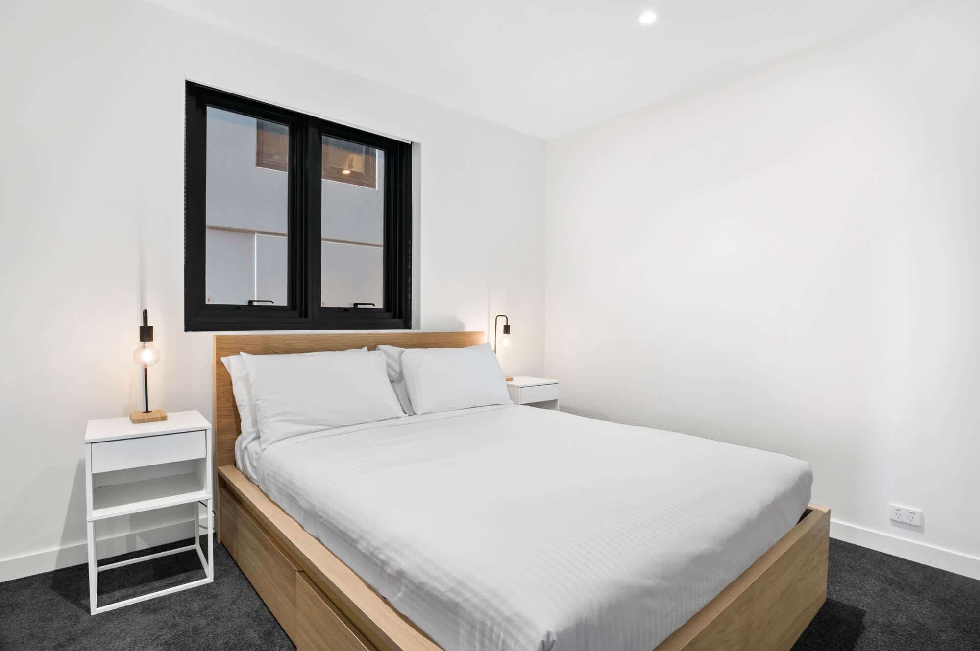 Un dormitorio moderno con un cabecero de madera, ropa de cama blanca, una mesita de noche con una lámpara y una ventana con marcos negros.