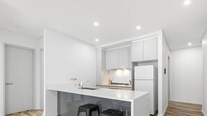 A modern kitchen with white cabinetry, a central island with stools, and a built-in oven. - New 2 bedroom apartment. Garden view with pool, jacuzzi,sauna & gym. (Eastgardens)