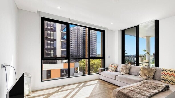 A modern living room with a large window, a sofa, a TV, and a dining area. - New 2 bedroom apartment. Garden view with pool, jacuzzi,sauna & gym. (Eastgardens)