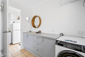 Une petite buanderie équipée d’un lave-linge, d’un évier, d’un miroir et d’une porte donnant sur une autre pièce.