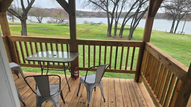 Eine HolzdTerrasse mit einem Glastisch und zwei Stühlen, mit Blick auf eine Wiese und ein Gewässer.