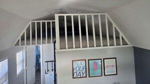 Ein Zimmer mit schrägem Giebeldach, einer weißen Treppe und einem Deckenventilator. An der Wand hängen drei gerahmte Bilder.