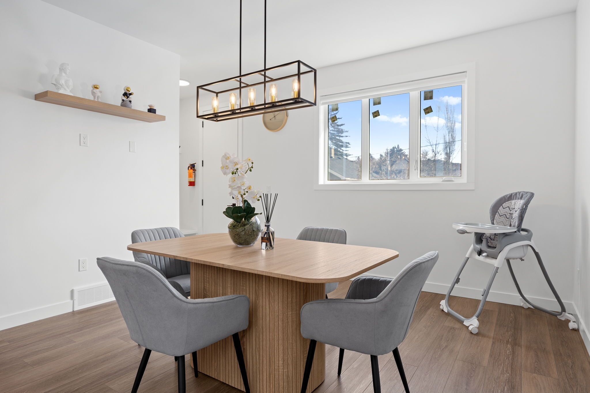 A modern dining area with a wooden table, grey chairs, a baby high chair, a hanging light fixture, and a window with a view of trees.