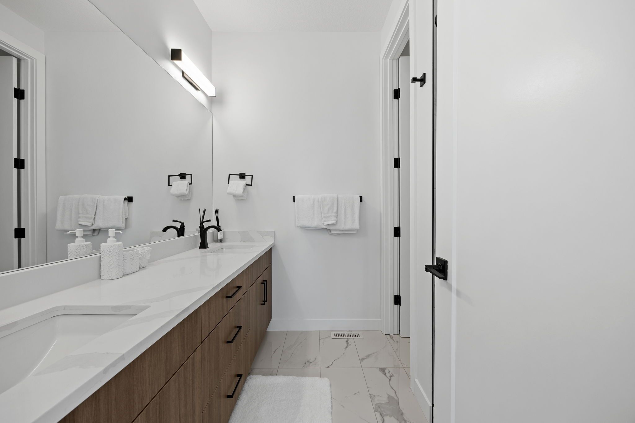 A modern bathroom with a large mirror, white sink, wooden cabinets, and a white floor.