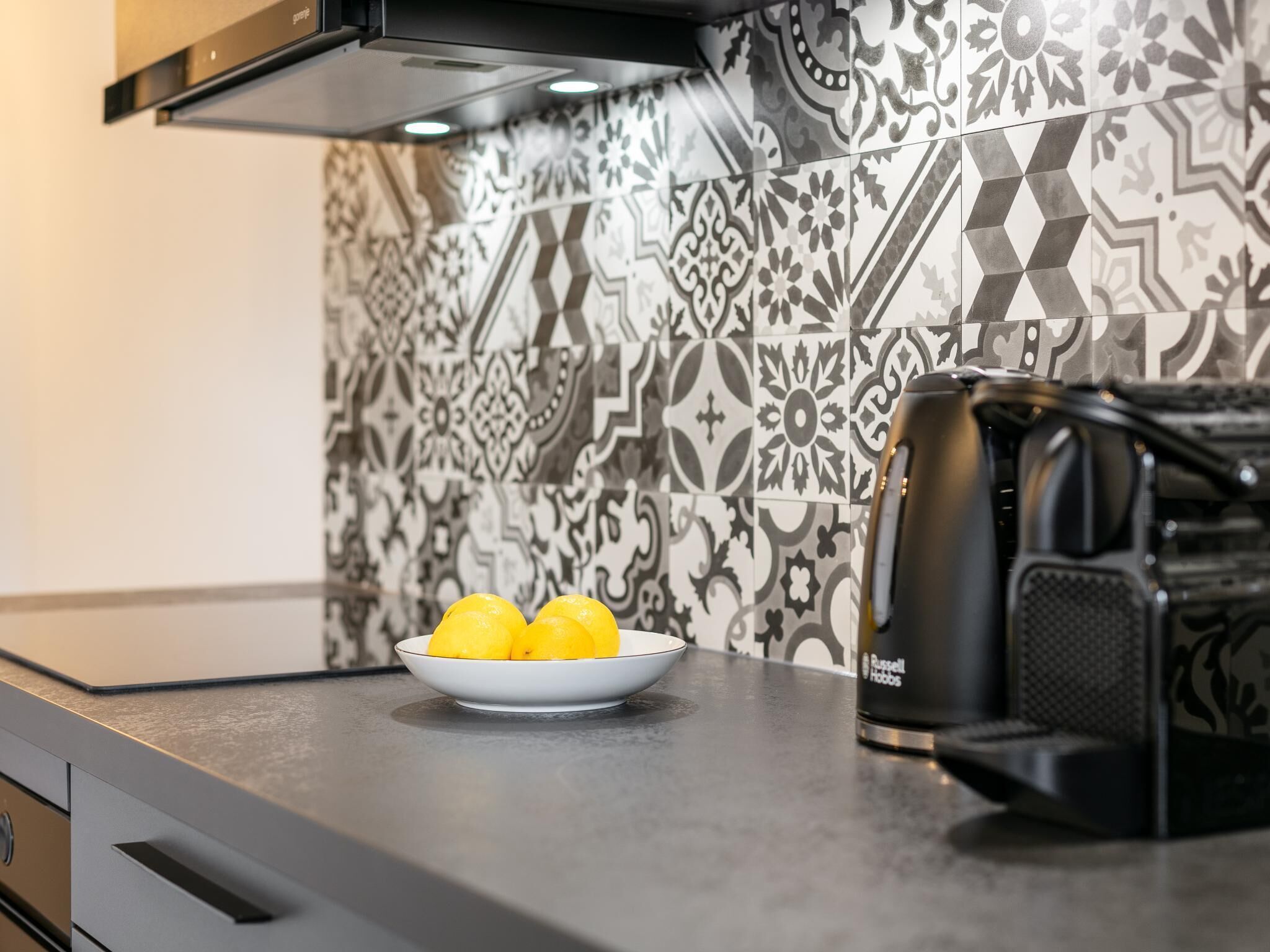 A kitchen with a patterned backsplash, a bowl of lemons, and a coffee maker.