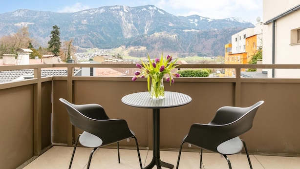 Ein Balkon mit zwei Stühlen, einem kleinen runden Tisch und einem Blumenstrauß. Im Hintergrund ist eine Bergsicht zu sehen.