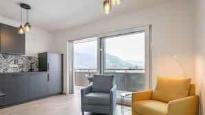Een modern appartement met keuken, grijze fauteuil, gele fauteuil, grote ramen en uitzicht op bergen.