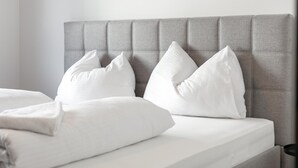 Een net opgemaakt bed met witte beddengoed en kussens, een grijs gestoffeerd hoofdbord, en een effen muur op de achtergrond.