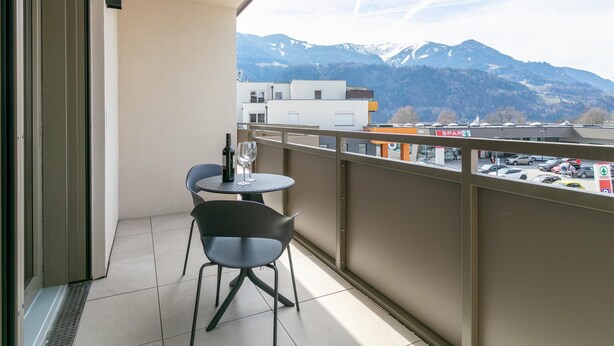 Een balkon met een tafel en stoelen, een fles wijn en uitzicht op bergen en een parkeerplaats.