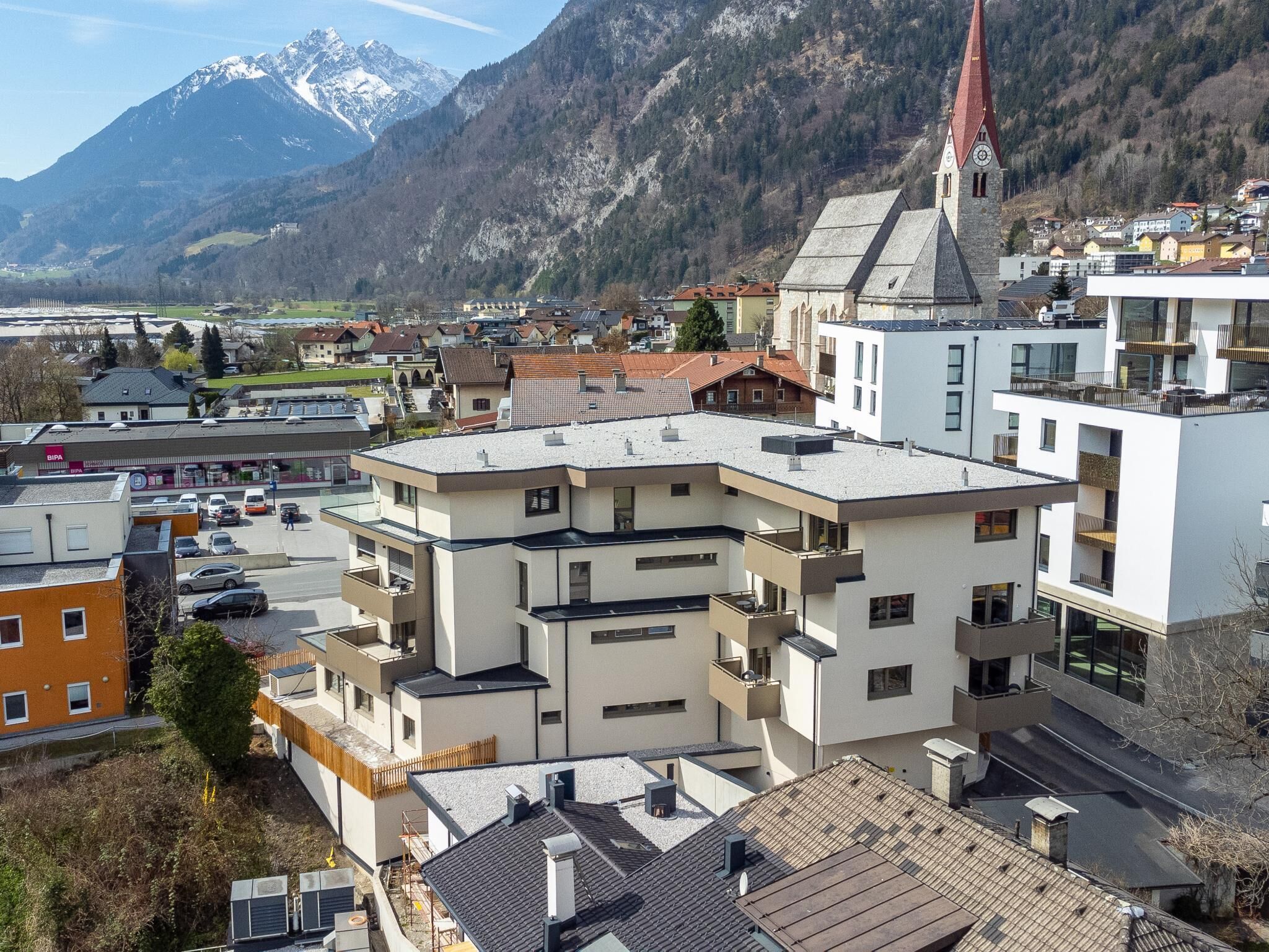 Ein mehrgeschossiges Gebäude mit flachem Dach, eine Kirche mit hohem Kirchturm und eine gebirgige Kulisse.