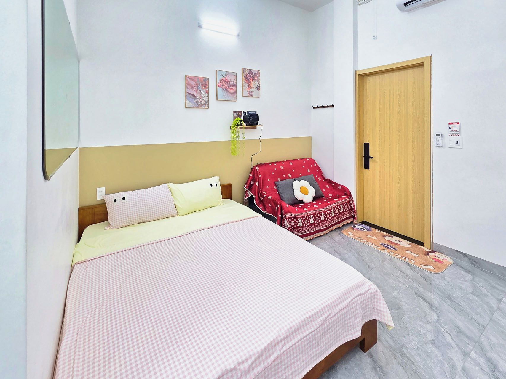 A bedroom with a bed, a red sofa, a wooden door, and a wall-mounted shelf with a plant.