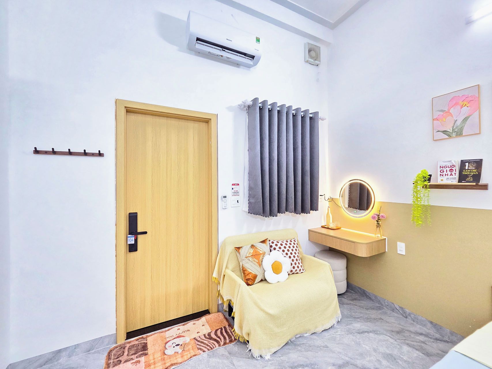 A room with a wooden door, an air conditioning unit, a bed with a patterned rug, a bedside table with a mirror, and a wall-mounted shelf with books and a plant.