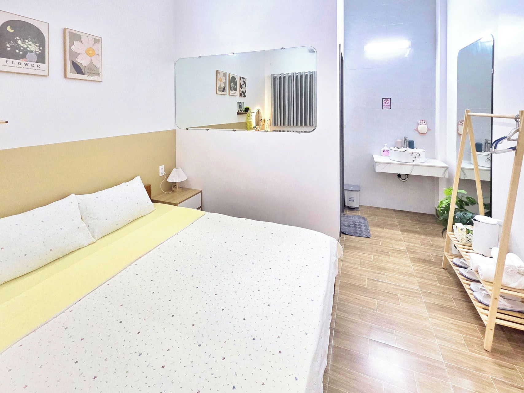 Habitación de hotel con cama, mesita de noche con lámpara, espejo, radiador, lavamanos y estante con toallas.