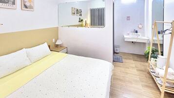 Habitación de hotel con cama, mesita de noche con lámpara, espejo, radiador, lavamanos y estante con toallas.