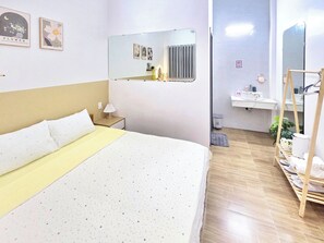 A hotel room with a bed, a bedside table with a lamp, a mirror, a radiator, a sink, and a shelf with towels.